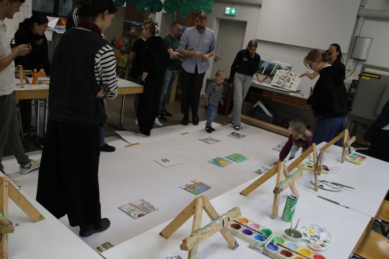 Papier liegt auf dem Boden, Menschen stehen drum herum und besprechen die Gestaltung des gemeinsamen Bildes