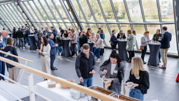 Gruppe von Personen beim Netzwerken in einem hellen, modernen Veranstaltungsraum im Wissenschaftspark Gelsenkirchen. Die Teilnehmer stehen an Stehtischen und tauschen sich aus, während einige sich am Buffet mit Speisen und Getränken versorgen. Der Raum ist mit großen Glasfronten ausgestattet, die den Blick auf die umliegende Landschaft freigeben.