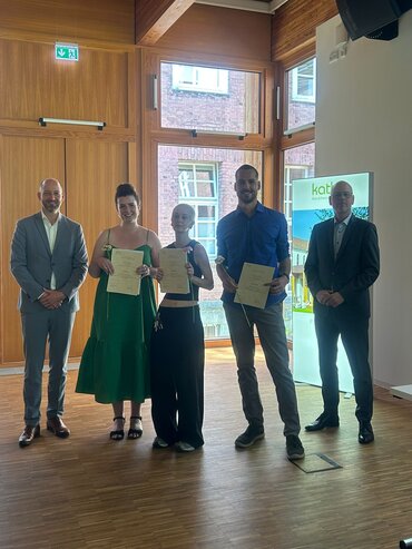 Fünf Personen sind auf dem Foto zusehen. Die drei besten Absolvent_innen zeigen Ihre Urkunden. Sie werden von zwei Mitarbeitenden der Pax-Bank flankiert, die das Preisgeld gestiftet hat.