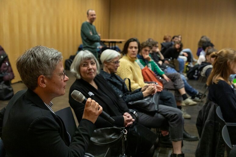 Eine Stuhlreihe eines Publikums. Im Vordergrund eine Frau die ein Mikrofon hält. Das restliche Publikum ist zu ihr gerichtet.