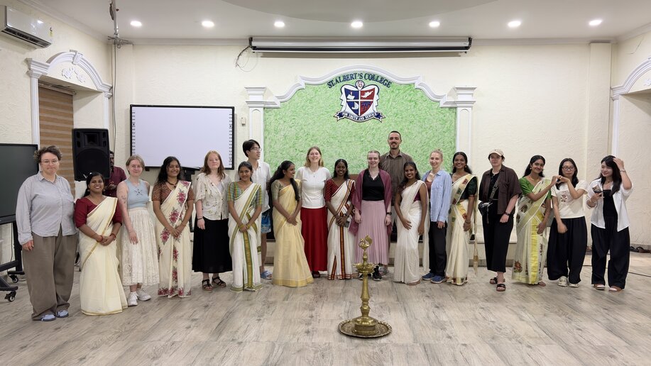 Gruppenfoto in einem Saal: 19 Personen in einer Reihe vor einer grünen Wand mit Wappen; vorne steht eine große traditionelle Messinglampe, einige tragen Sarees.