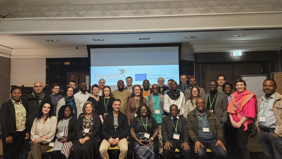 Eine Gruppe von Menschen posiert für ein Gruppenfoto in einem Konferenzraum. Alle tragen Namensschilder und einige haben grüne Lanyards. Im Hintergrund ist eine Projektionsleinwand mit dem Logo „MANDELA“ und dem Hinweis auf eine Co-Finanzierung durch die Europäische Union zu sehen. Die Gruppe lächelt und die Stimmung wirkt freundlich und zusammenhaltend.