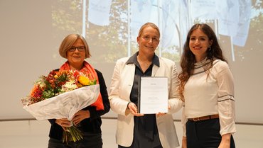 Drei Frauen stehen auf einer Bühne und lächeln freundlich in die Kamera. Ganz linkst steht Helene Hofmann, sie hält einen Blumenstrauß in der Hand, in der Mitte übergibt Julia Quirll da Matta die Urkunde an die rechts neben ihr stehende Angela Nehme.