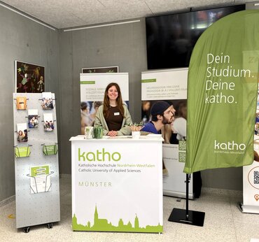 Eine Studentin steht lächelnd hinter der Theke des Messestandes der katho, Standort Münster. Drumherum sind zwei Roll-ups zu den Studiengängen Soziale Arbeit und Heilpädagogik zu sehen, ein Flyerständer und ein Beachflag mit der Aufschrift "Dein Studium. Deine katho".