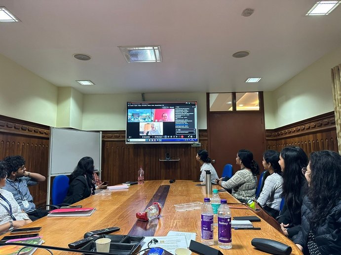 Indische Studierende der Christ University sitzen während einer Lehrveranstaltung an einem runden Tisch, im Hitnergrund ist eine Leinwand mit einer Videokonferenz zugeschalteter Seminarteilnehmener_innen.