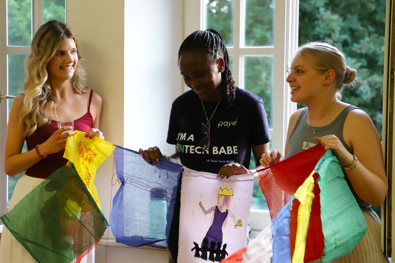 Drei Studentinnen stehen innen vor einem Fenster und halten eine Schnur mit mehreren bunten Flaggen. Auf der mittleren ist eine Frau mit Krone zu sehen, vor ihren Füßen stehen kleine schwarze Figuren.