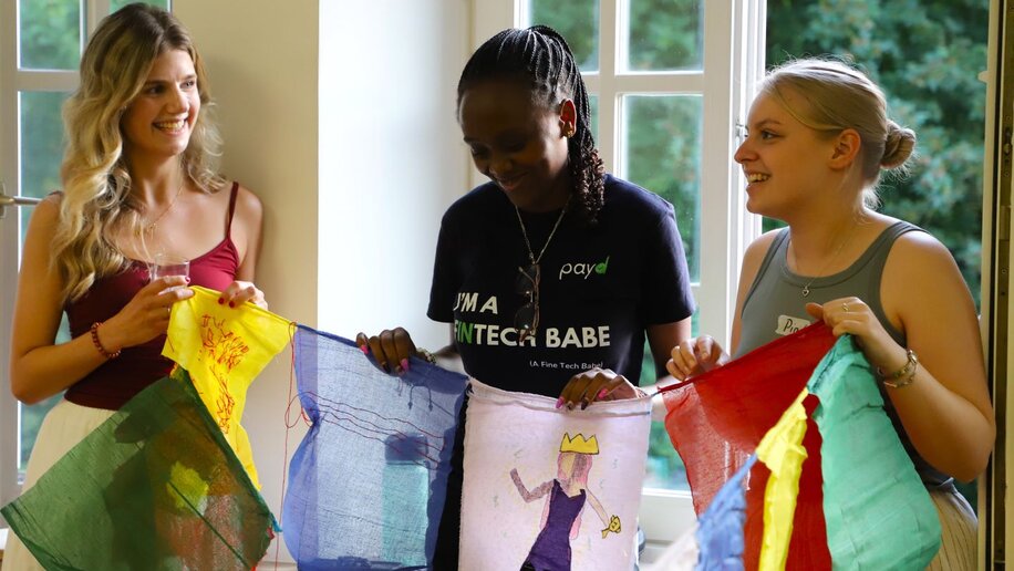 Drei Studentinnen stehen innen vor einem Fenster und halten eine Schnur mit mehreren bunten Flaggen. Auf der mittleren ist eine Frau mit Krone zu sehen, vor ihren Füßen stehen kleine schwarze Figuren.