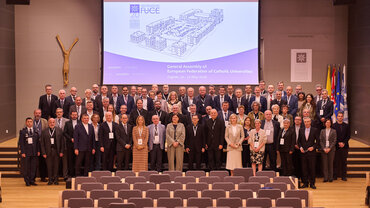 Gruppenbild mit zahlreichen festlich gekleideten Personen in einem großen Saal. Über den Personen ist auf einer Leinwand " General Assembly of European Federation of Catholic Universities" zu lesen.