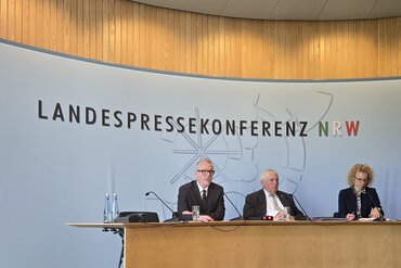 Michael Isfort, Karl-Josef-Laumann und eine weitere Person sitzen an einem großen Redepult. Auf der Wand dahinter steht ein großer Schriftzug "Landespressekonferenz NRW".