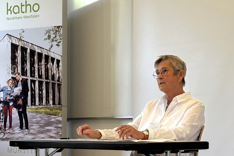 Professorin Tölle sitzt an einem Tisch und spricht. Neben ihr steht ein katho-Roll-up.