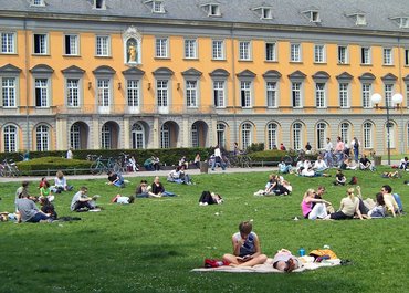 Studierende sitzen im Hofgarten auf einer grünen Wiese im Sommer vor dem Bonner Universitätsgebäude.