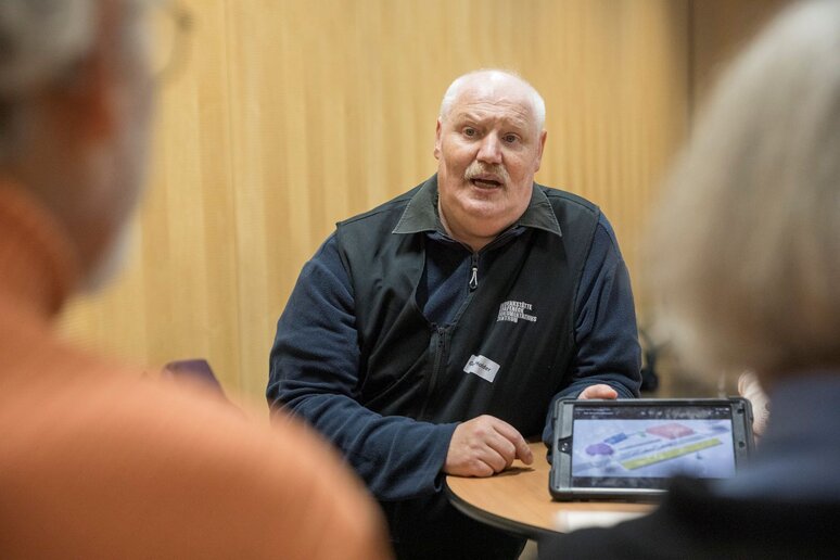 Ein älterer Mitarbeiter der Gedenkstätte Grafeneck zeigt zwei Besuchern eine Grafik auf einem Tablet. Der Mann hat weiße Haare und trägt schwarze Kleidung mit dem Gedenkstätten Grafeneck Logo.