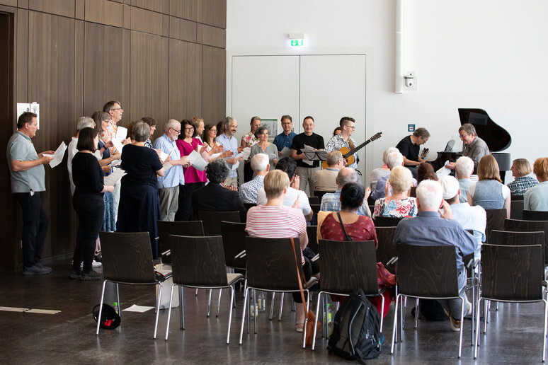 Viele Personen stehen vor- und hintereinander und singen. Unterstützung erhält der Chor durch einen Gitarren- und einen Klavierspieler. Das Publikum hört interessiert zu.