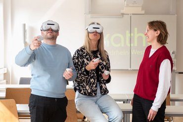 Zwei Personen in einem Seminarraum tragen VR-Brillen, eine weitere beobachtet die beiden.