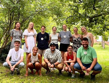 Die Gruppe der Zertifizierten präsentiert sich in zwei Reihen auf einer Rasenfläche, vor einem Blätterwald.