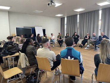 Das Bild zeigt Menschen, die im Stuhlkreis in einem Raum sitzen und miteinander reden.
