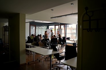 Mehrere Menschen sitzen in einem Stuhlkreis. Zwei Personen stehen vor einer Flipchart und sind im Gespräch