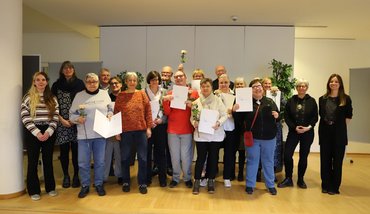 Gruppenbild mit 18 Personen. Die stolzen Kursteilnehmer_innen halten Urkunden und helle Rosen in der Hand.