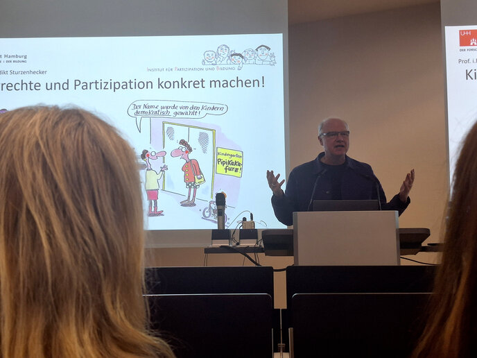 Prof. i. R. Dr. Benedikt Sturzenhecker steht am Rederpult und erklärt, wie man Kinderrechte und Partizipation in Einrichtungen konkret macht.