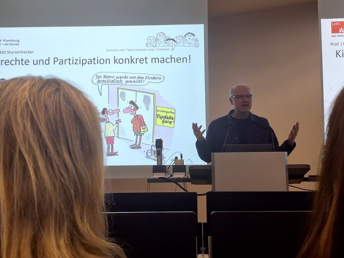Prof. i. R. Dr. Benedikt Sturzenhecker steht am Rederpult und erklärt, wie man Kinderrechte und Partizipation in Einrichtungen konkret macht.