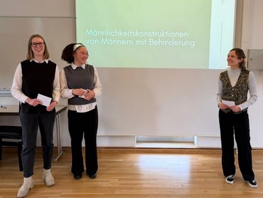 Eine Folie der PPT wird an die Wand geworfen; drei Studierende stehen rechts und links daneben.