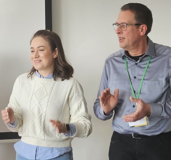 Zwei Personen stehen in einem Raum und scheinen in Bewegung oder beim Applaudieren zu sein. Die Frau trägt einen hellen Pullover und eine blaue Bluse, der Mann ein Hemd mit grünem Lanyard. Beide lächeln und schauen in dieselbe Richtung, die Atmosphäre wirkt fröhlich und motiviert.