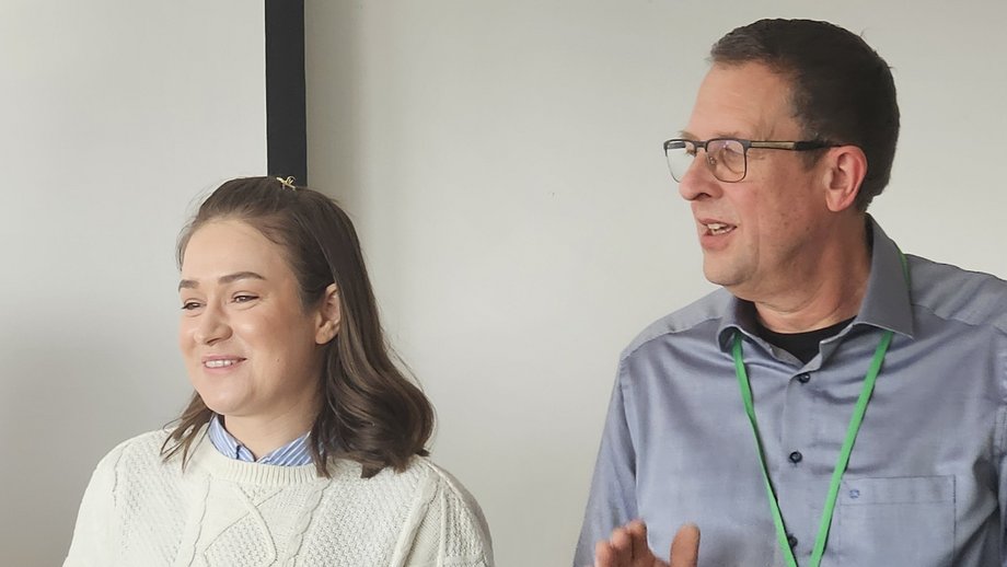 Zwei Personen stehen in einem Raum und scheinen in Bewegung oder beim Applaudieren zu sein. Die Frau trägt einen hellen Pullover und eine blaue Bluse, der Mann ein Hemd mit grünem Lanyard. Beide lächeln und schauen in dieselbe Richtung, die Atmosphäre wirkt fröhlich und motiviert.