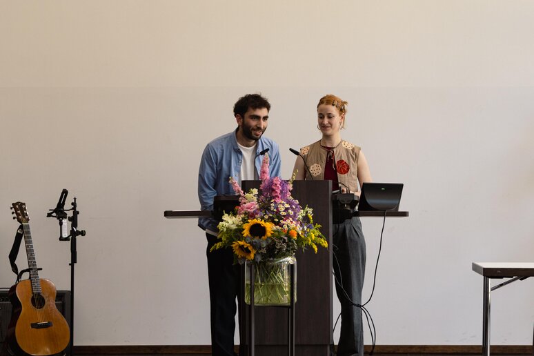 Zwei Studierende halten am Rednerpult auf der Bühne eine Rede. Die Bühne ist dekoriert mit einem schönen, ausladenden Blumenstrauß.