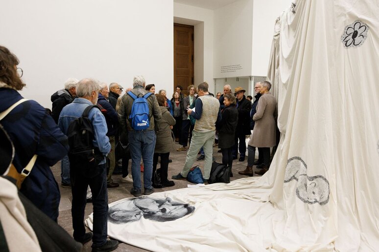 Eine große Gruppe Menschen steht vor einem Ausstellungsstück. Das Ausstellungsstück ist ein großes weißes Tuch, auf dem verschiedene schwarz-weiß Abbildungen von Kindern aufgenäht wurden.