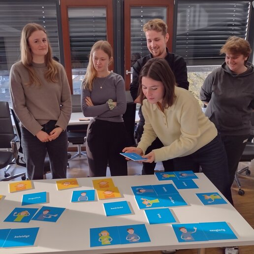 Schulungsteilnehmende stehen um einen Tisch mit blauen Karten, auf denen Begriffe wie neugierig oder ängstlich stehen.