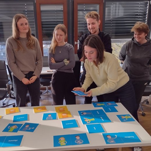 Schulungsteilnehmende stehen um einen Tisch mit blauen Karten, auf denen Begriffe wie neugierig oder ängstlich stehen.