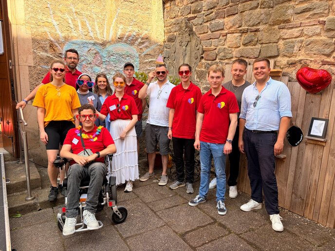 Zwölf junge Menschen stehen für ein Gruppenbild vor einer Wand. Viele von ihnen tragen ein rotes Poloshirt, eine Frau trägt Nonnentracht und eine Sonnenbrille mit Herzen, eine Person sitzt im Rollstuhl.