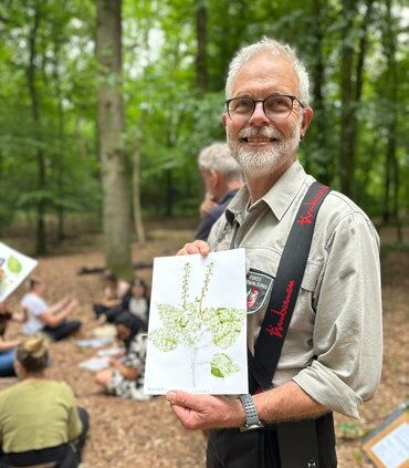 Ein Mann in Arbeitskleidung hält ein Blatt Papier in der Hand, auf dem eine Pflanze gezeichnet ist. Im Hintergrund sind Menschen im Wald zu sehen.