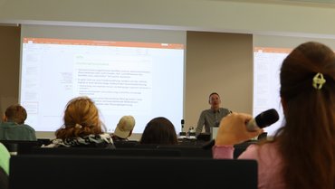 Ein Professor beim Vortrag vor Zuschauerschaft im Hörsaal, im Vordergrund des Bildes Rückansicht einer Person mit Handmikrofon