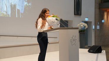 Angela Nehme während ihrer Rede beim Rathausempfang in Köln. Sie steht auf der Bühne, das Foto ist seitlich von ihr hinter dem Rednerpult aufgenommen. Sie hält einen Blumenstrauß in der Hand und blickt geradeaus ins Publikum.