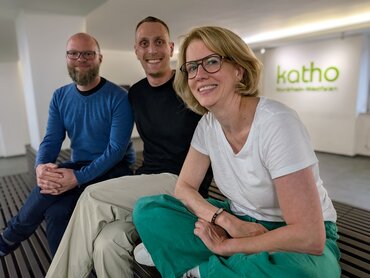 Zwei Männer und eine Frau sitzen auf einer Bank, im Hintergrund leuchtet an der Wand das katho-Logo