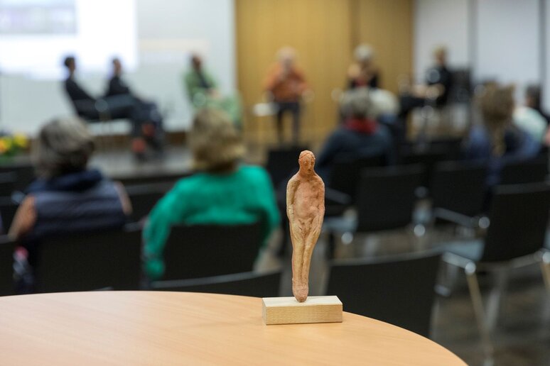 Eine kleine Tonfigur eines nackten Mannes steht auf einem Tisch. Die Figur ist im Fokus, im Hintergrund sind eine Bühne und Publikum zu erkennen.