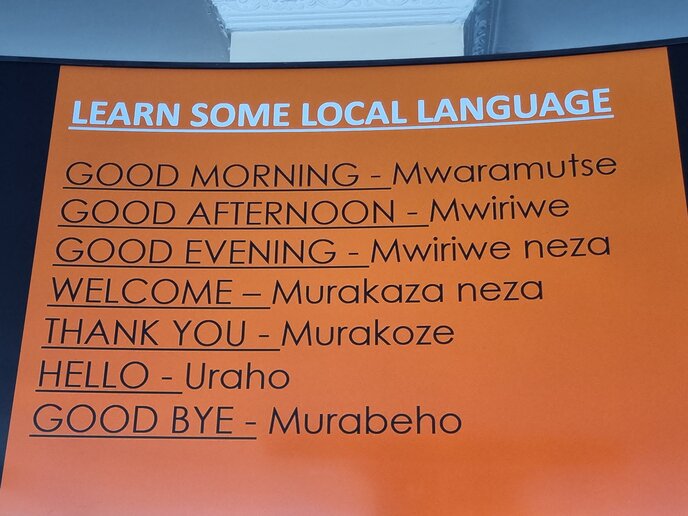 Ein Plakat mit der Überschrift "Learn some local language". Darunter sind die Übersetzungen in der lokalen Sprache aufgeführt für: Good morning, good afternoon, good evening, welcome, thank you, hello, goodbye.