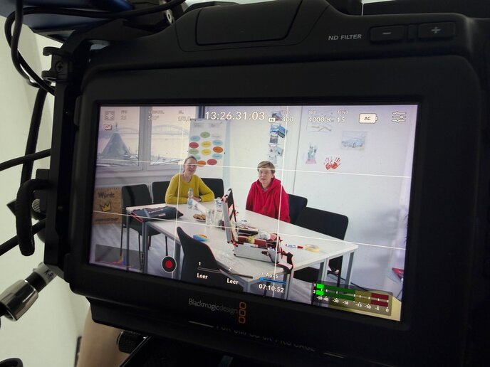 Zwei Personen sitzen nebeneinander an einem Tisch und werden durch die Linse der Filmkamera fotografiert.