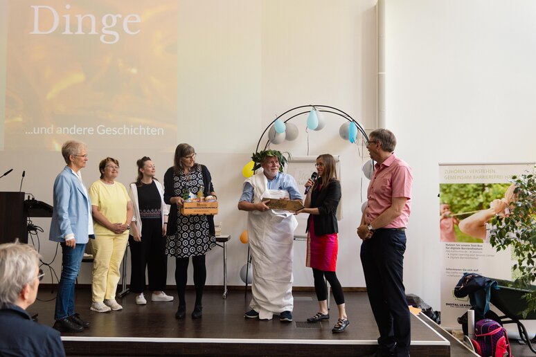 Der geehrte Kollege steht im Kreise seiner Kolleg_innen, verkleidet mit einem Blätterkranz auf dem Kopf und in eine weiße Tischdecke gehüllt.