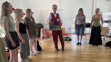 Fünf junge Frauen mit einer roten Clownsnase stehen im Kreis. In der Mitte der Klinik-Clown, gekleidet mit einem karierten Anzug. 