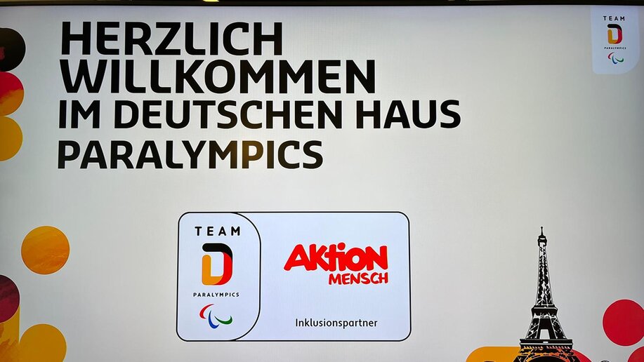 PRojektion an die Wand mit der Aufschrift: Herlich willkommen im Deutschen Haus Paralympics. Darunter befindet sich das Logo der Aktion Mensch und Team Deutschland