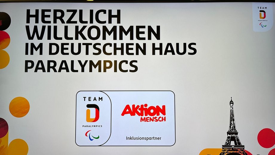 PRojektion an die Wand mit der Aufschrift: Herlich willkommen im Deutschen Haus Paralympics. Darunter befindet sich das Logo der Aktion Mensch und Team Deutschland