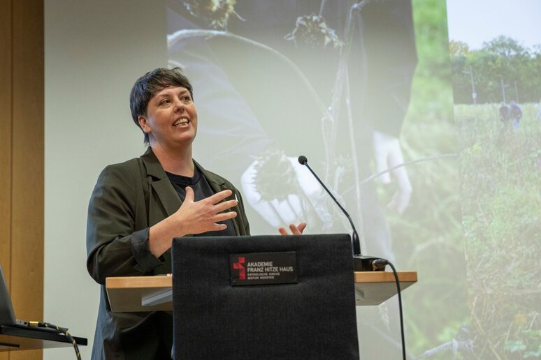 Eine Frau steht am Rednerpult. Sie hat kurze dunkle Haare und trägt einen Blazer. Im Hintergrund ist eine Präsentation zu erkennen.