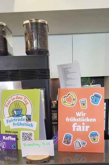 Infoschilder, die das Fairtrade-Frühstück am 13.5. auf einem grünen und einem orangefarbenen Schild bewerben.