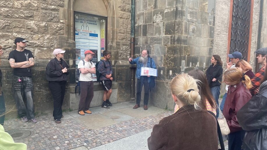 Eine Gruppe von Personen steht im Halbkreis vor einer steinernen Kirchenfassade; eine Person hält ein Blatt und spricht, die anderen hören zu.