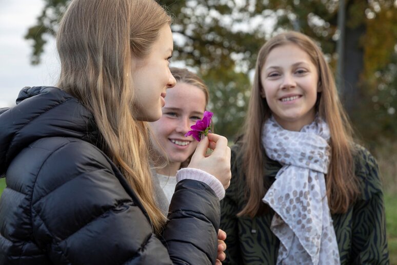 Drei Schülerinnen stehen auf einer Wiese. Eine hält eine lila Blume an der sie riecht.