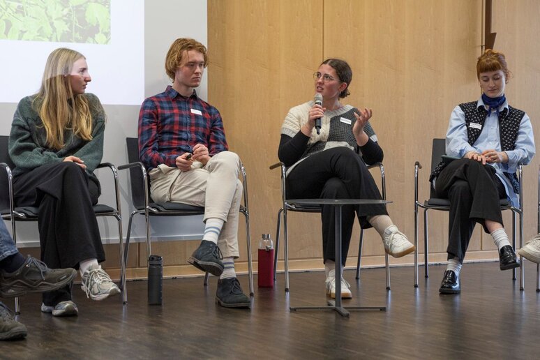 Vier junge Menschen sitzen auf dem Podium vor einer Präsentation. Eine junge Frau hält ein Mikrofon und spricht.