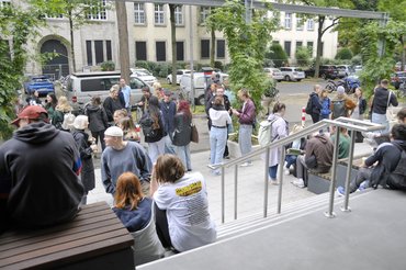 Studierende sitzen in der Pause auf den Treppen vor dem Eingang zur Hochschule. 