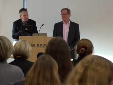 Zwei Männer im Jacket stehen an einem Redepult. Im Vordergrund sitzt Publikum.